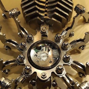 May include: A silver metal sculpture of a spider with a clock face in the center. The spider has eight legs and a body made of many small metal pieces.