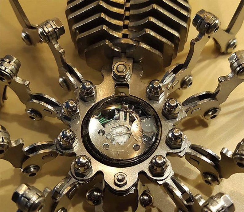 May include: A silver metal sculpture of a spider with a clock face in the center. The spider has eight legs and a body made of many small metal pieces.