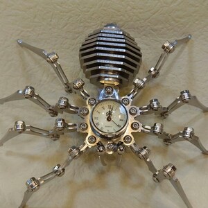 May include: A silver metal spider clock with a round clock face and black Roman numeral hour markers. The clock face says "Quartz".