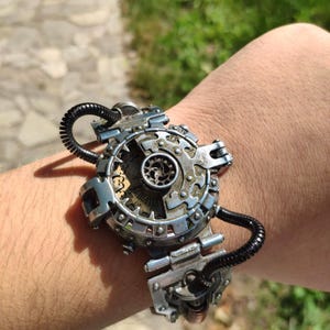Op de afbeelding: Een zilveren steampunk-armband met een cirkelvormig tandwielontwerp en zwarte slangaccenten.