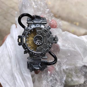 Op de afbeelding: Een zilveren steampunk-stijl polshorloge met een ronde wijzerplaat en Romeinse cijfers. Het horloge heeft een complex ontwerp met tandwielen en pijpen.