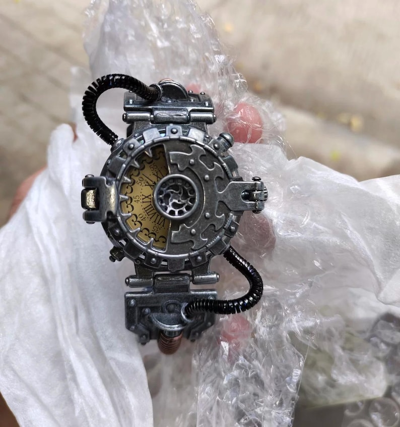 Op de afbeelding: Een zilveren steampunk-stijl polshorloge met een ronde wijzerplaat en Romeinse cijfers. Het horloge heeft een complex ontwerp met tandwielen en pijpen.