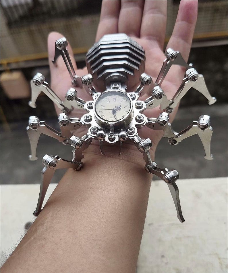 May include: A silver metal spider clock with a round clock face and black hands. The clock is on a person's wrist.