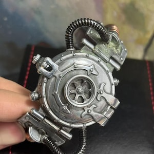 May include: A silver steampunk-style bracelet with a round, detailed, gear-like design. The bracelet has a black, flexible tube attached to it.