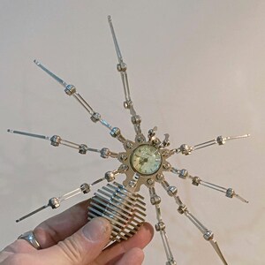 May include: A silver metal spider sculpture with eight articulated legs and a clock face in the center. The clock face has black Roman numeral hour markers and black hands.