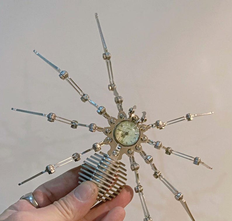 May include: A silver metal spider sculpture with eight articulated legs and a clock face in the center. The clock face has black Roman numeral hour markers and black hands.