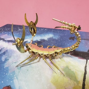 May include: A gold and copper colored metal scorpion sculpture with a steampunk design. The scorpion is posed with its tail raised and its claws extended. The sculpture is on a blue and white background.