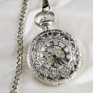 Op de afbeelding: Een zilveren zakhorloge met een ingewikkeld, sierlijk ontwerp en een skeletwijzerplaat. Het horloge heeft Romeinse cijfers voor de uren en een ketting.