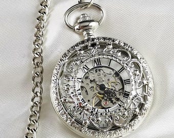 May include: A silver pocket watch with an intricate, ornate design and a skeletonized face. The watch has Roman numeral hour markers and a chain attached.