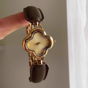 Peut inclure: Montre-bracelet dorée avec un cadran en forme de trèfle et un bracelet à maillons dorés. La montre présente un cadran doré texturé et des bracelets en cuir marron. La montre est tenue par un doigt.