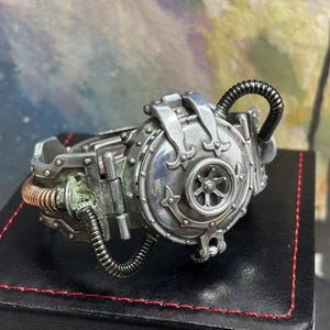 May include: A silver steampunk-style bracelet with a round, detailed, gear-like design. The bracelet has a copper-colored pipe and a black pipe attached to it.