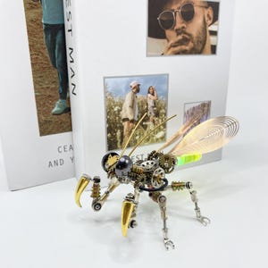 May include: A steampunk-style insect sculpture made of metal gears and wires. The insect has gold-colored legs and antennae, and its wings are made of a translucent, iridescent material.