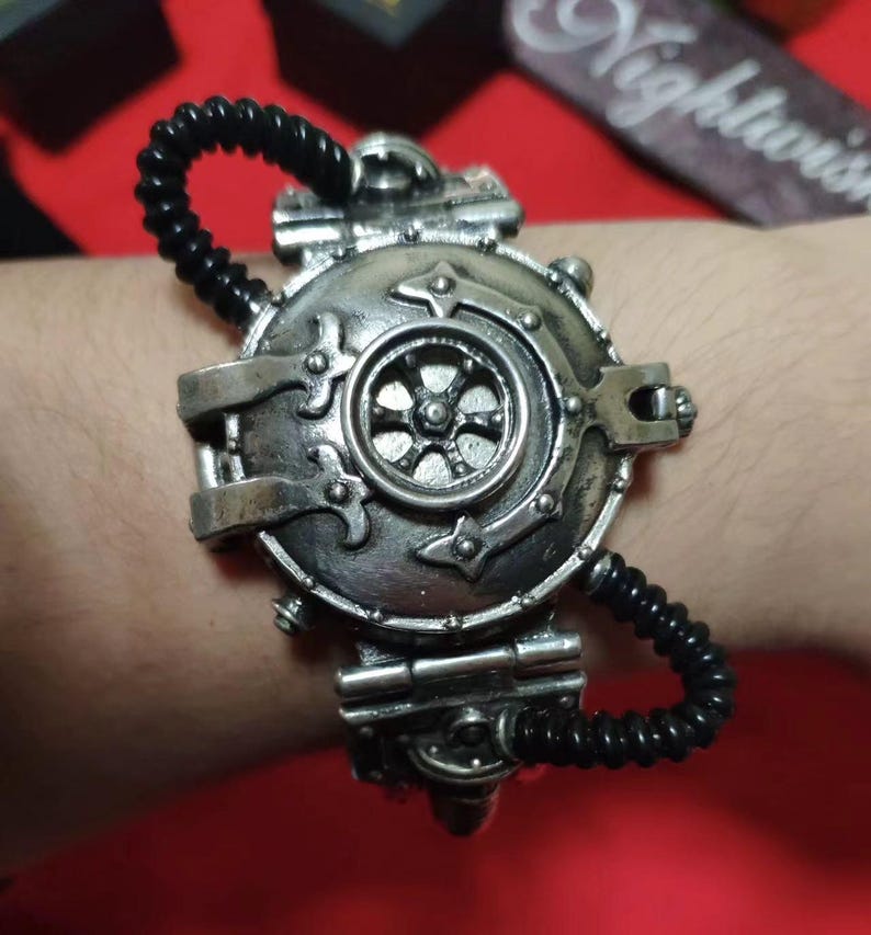 May include: A silver steampunk-style bracelet with a round, detailed, gear-shaped charm. The bracelet is made of metal and has a black elastic band.