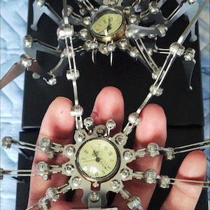 May include: Two silver metal spider clock sculptures. The first spider is on a black surface and has a round clock face with Roman numerals. The second spider is held in a hand and has a round clock face with Arabic numerals.