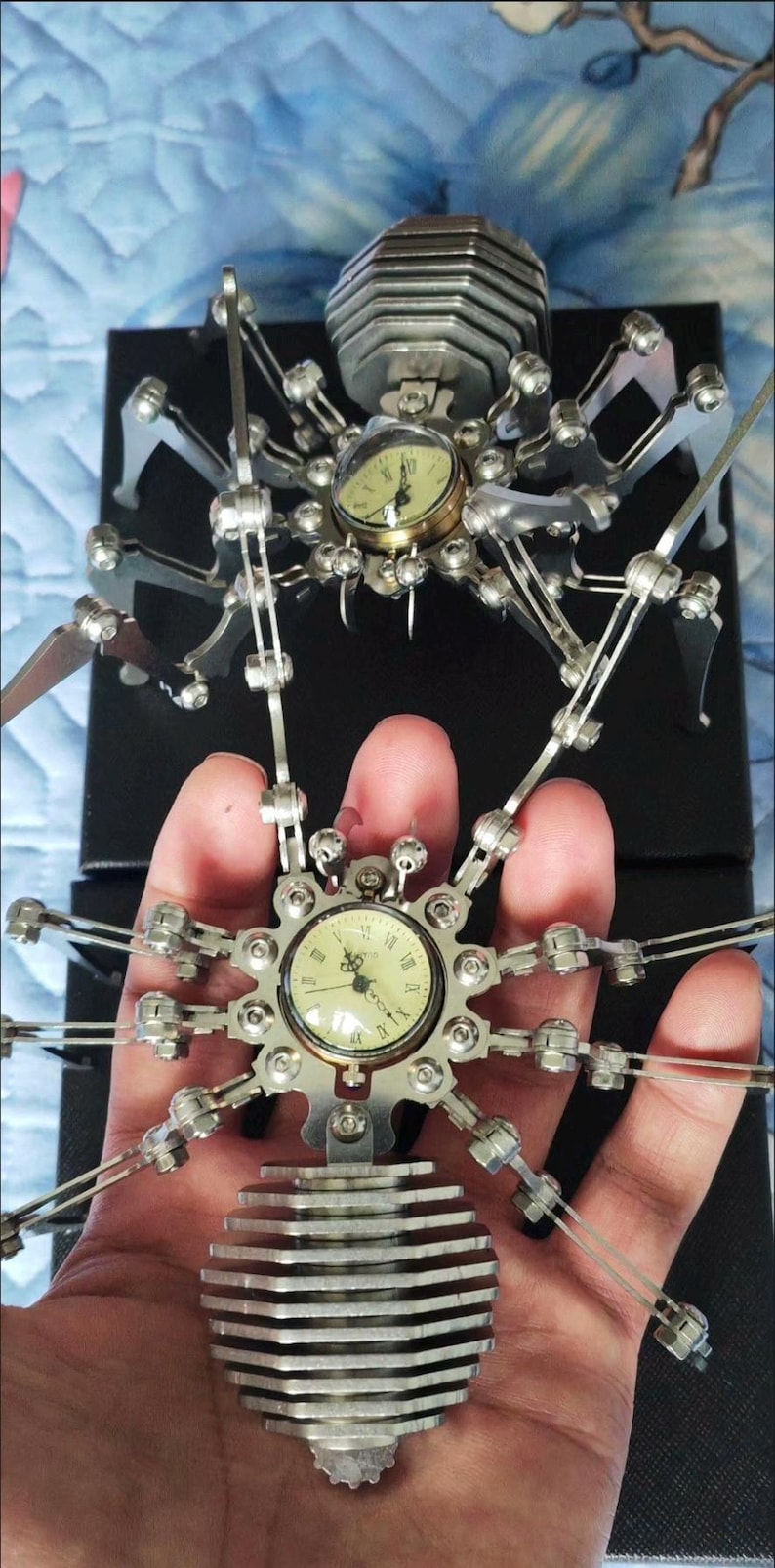 May include: Two silver metal spider clock sculptures. The first spider is on a black surface and has a round clock face with Roman numerals. The second spider is held in a hand and has a round clock face with Arabic numerals.