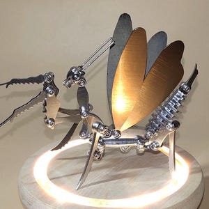 May include: A metal praying mantis sculpture with articulated legs and wings. The wings are made of a shiny, gold-coloured metal. The sculpture is mounted on a wooden base with a glowing ring of light.