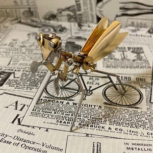 May include: A steampunk metal dragonfly sculpture with brass wings and intricate mechanical details. The dragonfly is made of silver and brass-coloured metal, set against a vintage newspaper background.