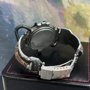 May include: A silver and copper steampunk-style wristwatch with a round face and a detailed, intricate design. The watch has a unique, adjustable band with a buckle closure.