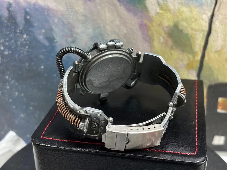 May include: A silver and copper steampunk-style wristwatch with a round face and a detailed, intricate design. The watch has a unique, adjustable band with a buckle closure.