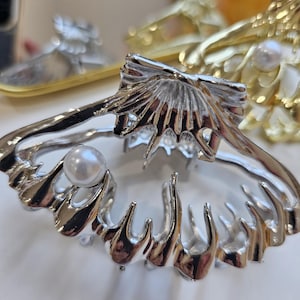 Könnte beinhalten: Eine silberfarbene Haarspange in Form einer Muschel, mit einem Perlenakzent. Die Spange hat ein detailliertes, strukturiertes Design und ist vor einem weichen, neutralen Hintergrund platziert. Weitere ähnliche Haaraccessoires sind im Hintergrund zu sehen.