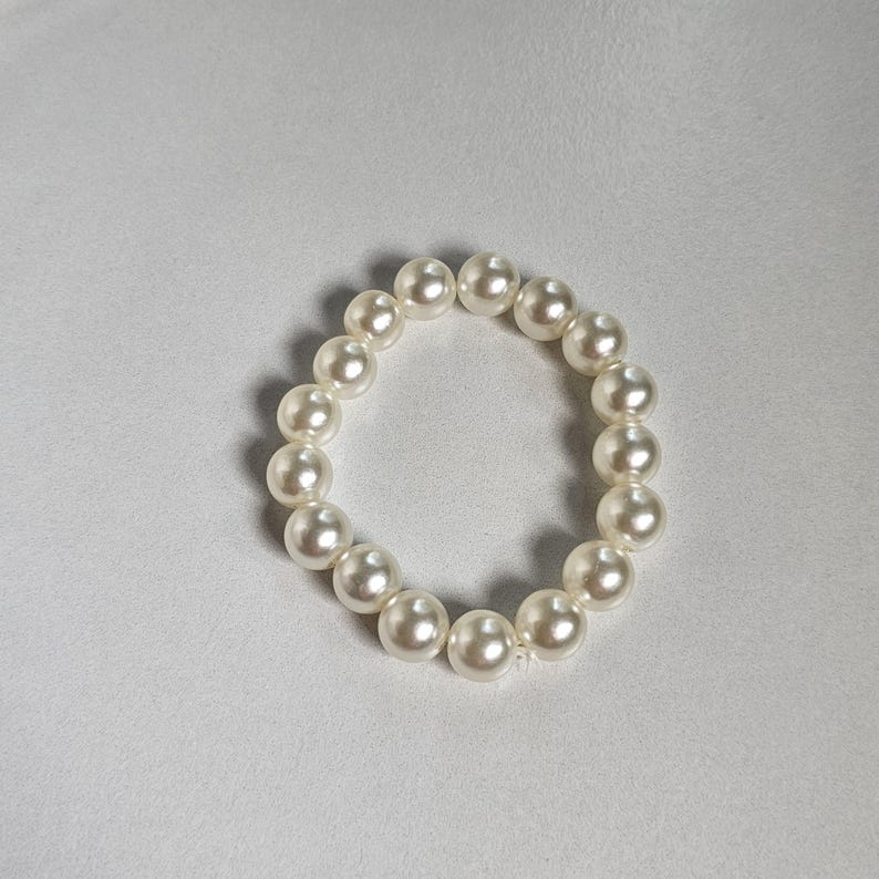 May include: A round, off-white pearl bracelet. The pearls are evenly sized and strung together, forming a complete circle. The bracelet is set against a plain, light-colored background.