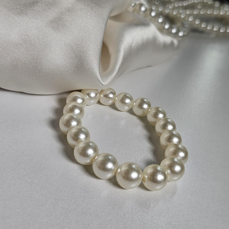 May include: A cream-colored pearl bracelet with large, round beads. The bracelet is displayed on a white satin surface, with a strand of smaller pearls visible in the background. The pearls have a lustrous sheen.