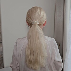 May include: Blonde hair styled in a low ponytail, secured with a pearl hair tie. The hair tie is made of small, round, white pearls. The person is wearing a light pink blouse.