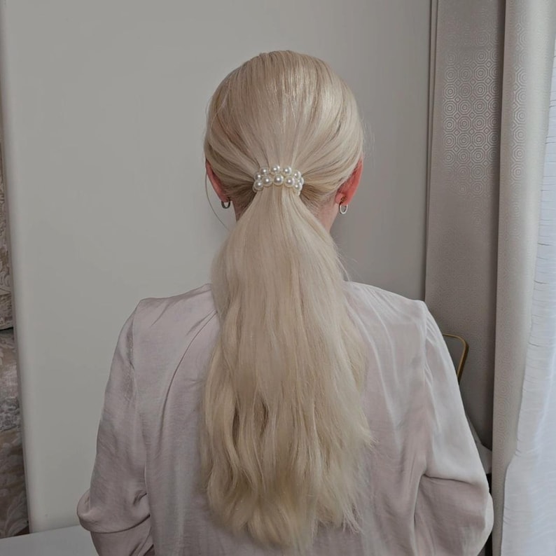 May include: Blonde hair styled in a low ponytail, secured with a pearl hair tie. The hair tie is made of small, round, white pearls. The person is wearing a light pink blouse.