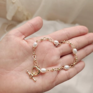 May include: A gold-toned chain bracelet featuring oval white pearls interspersed with textured oval links. The bracelet includes a heart-shaped toggle clasp closure, which serves as the focal point of the piece, connecting the delicate chain segments.