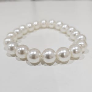 May include: A white pearl bracelet with large, round beads. The beads are strung together on an elastic band, creating a simple yet elegant accessory. The bracelet is shown on a white surface.