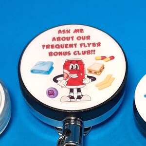 May include: Three white retractable badge holders with a black base. The badge holders have a cartoon image of a can of soda with a smiling face and the text "Ask me about our frequent flyer bonus club!" printed on them.