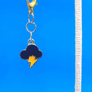May include: A pair of earrings with a gold chain and a black cloud with a yellow lightning bolt charm. The earrings are displayed on a white fabric stand against a blue background.