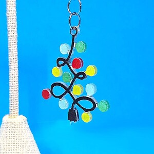 May include: A Christmas tree charm with a black outline and colorful lights. The charm is attached to a silver chain and clasp. The tree has red, yellow, and blue lights. The charm is displayed on a white stand against a blue background.