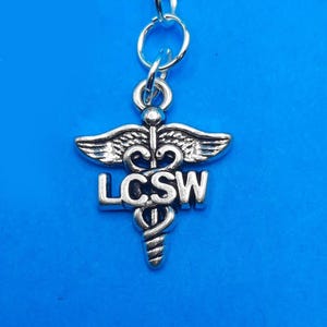 May include: A silver-colored charm with a lobster clasp. The charm features the caduceus symbol with the letters LCSW. The charm is suspended from a small ring and a larger clasp, all in silver.