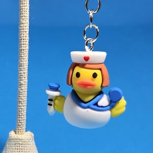 May include: A novelty charm featuring a nurse duck wearing a white uniform, blue collar, and a white cap with a red heart. The duck holds a syringe and a blue medical tool. The charm is attached to a silver-colored clasp.