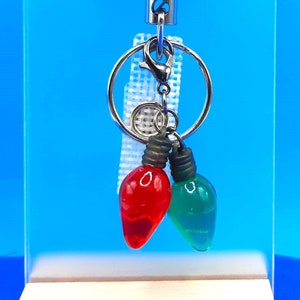 May include: A retractable badge reel with a Christmas theme. The reel features a circular design with an elf illustration and the text "So Elfin' Cute." Two miniature light bulb charms, one red and one green, dangle below.