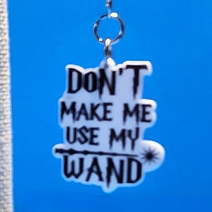 May include: A white acrylic charm with black text that reads DON'T MAKE ME USE MY WAND, featuring a small wand graphic underlined beneath the word USE. The charm is attached to a silver-toned metal lobster claw clasp by two small jump rings.