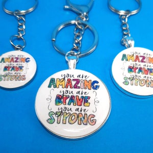 May include: Three silver keychains with round, white, silver-rimmed charms. Each charm has colorful text that reads "You are Amazing, You are Brave, You are Strong".