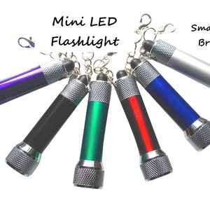 May include: Six mini LED keychain flashlights in different colors: purple, black, green, red, blue, and silver. The flashlights have a silver metal head and a keychain ring.