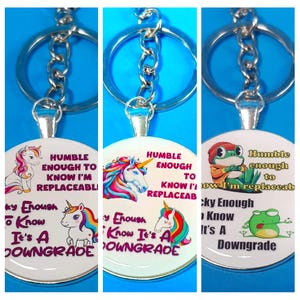 May include: Three keychains with round, white, and silver charms. Each charm features a different cartoon animal and text that reads "Humble enough to know I'm replaceable" and "Lucky Enough to Know It's a Downgrade".
