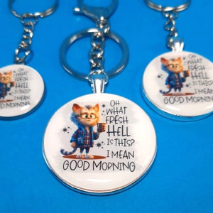 May include: Three silver keychains with round, white, printed charms featuring a cartoon cat holding a cup of coffee. The charms have the text "Oh what fresh hell is this? I mean good morning."