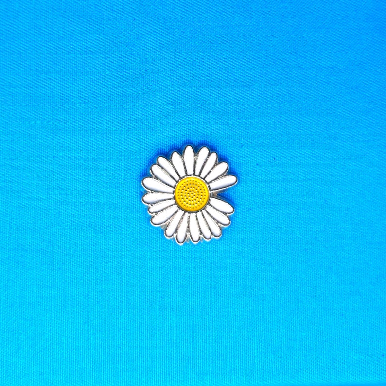 May include: A white daisy pin with a yellow centre on a blue background.