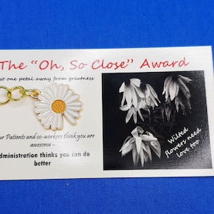 May include: A white card with the text "The "Oh, So Close" Award" and "Just one petal away from greatness". A gold chain with a white daisy charm is attached to the card. The card also has a black and white photo of wilted flowers with the text "Wilted flowers need love too".