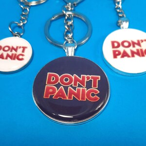 May include: Three silver keychains with round, white and blue enamel charms. Each charm features the text "Don't Panic" in red letters.