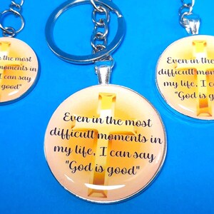 May include: Three circular silver-toned keychains featuring a gold cross graphic on a light orange background. Each keychain displays the black text: Even in the most difficult moments in my life, I can say God is good. The keychains have metal rings.