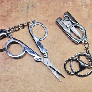 May include: Two small, silver-colored scissors on keychains. One pair is open, and the other is closed. The scissors are connected to a chain and a key ring. The background is a textured, tan surface.