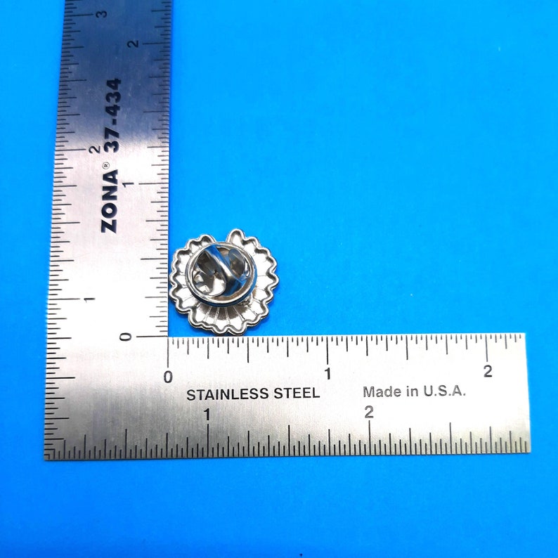 May include: A silver pin featuring a detailed design of a flower with a scalloped edge. The pin measures approximately 2.5 cm in diameter and has a safety clasp on the back.
