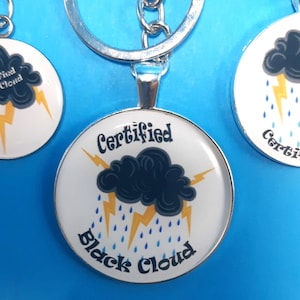 May include: A silver keychain with a round charm featuring a black cloud with lightning bolts and rain drops. The charm has the text "Certified Black Cloud" on it.