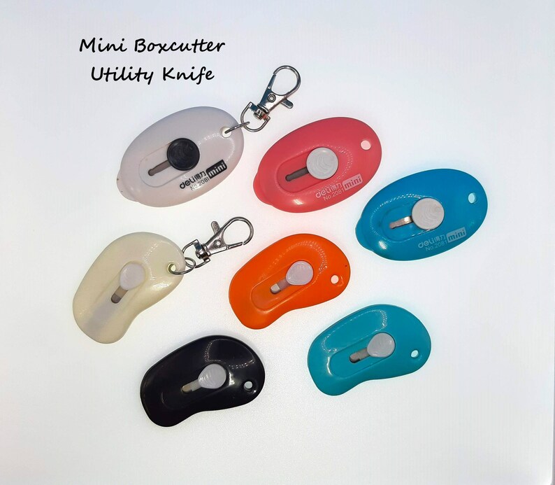 May include: Six colorful mini box cutters with keychains. The box cutters are shaped like a teardrop and have a silver blade. The text "OLFA mini" is printed on each box cutter.