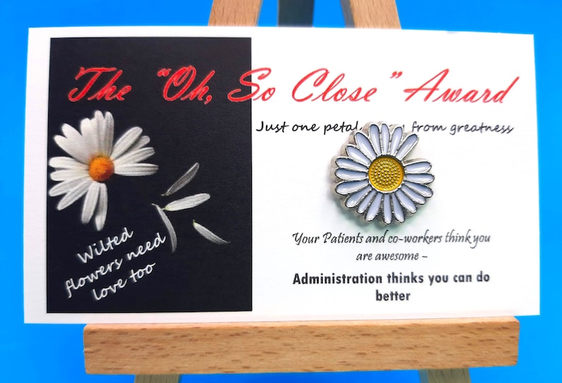 May include: A white card with black text that reads "The "Oh, So Close" Award" and "Just one petal, from greatness". The card also has a white daisy with a yellow centre on the right side. The card is on a small wooden easel.